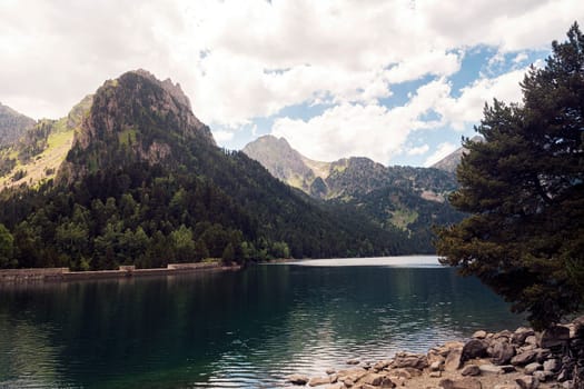spectacular landscape of calm lake surrounded by massive green mountains and coniferous trees against cloudy sky in Aiguestortes i Estany of Saint Maurici National Park