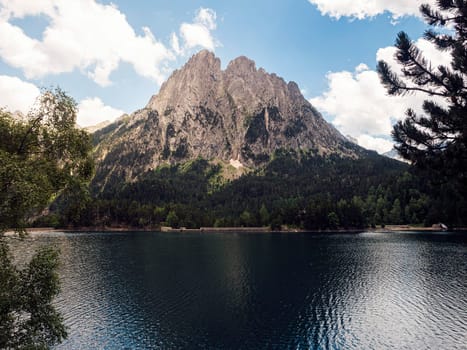 spectacular landscape of calm lake surrounded by massive green mountains and coniferous trees against blue sky in Aiguestortes i Estany of Saint Maurici National Park