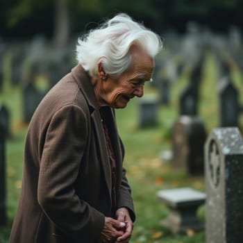 An elderly woman with white hair, wearing a brown coat, stands pensively before a gravestone in a cemetery, surrounded by other markers and greenery.