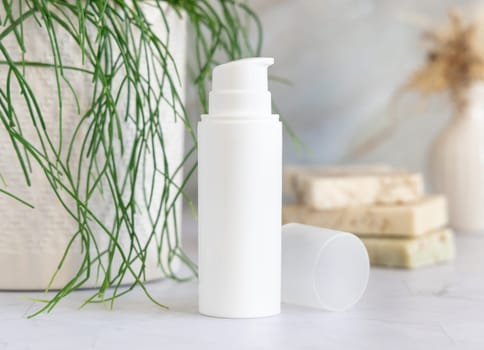 White cream pump bottle on white marble countertop near potted green plant in bathroom, close up, packaging mockup. Natural cosmetic product for face or body care