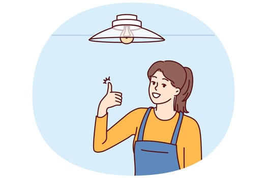 Woman electrician showing thumbs up after fixing chandelier or replacing light bulb under ceiling. Girl in overalls works as electrician efficiently performing tasks of setting up electricity