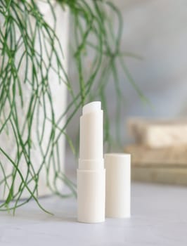 Opened lip balm stick on white marble conutertop near potted green plant in bathroom closeup, mockup. Lifestile scene with beauty product, everyday skincare routine