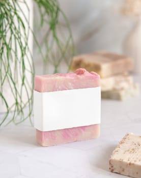 Pink soap bar with blank label on white marble countertop near potted green plant in bathroom, close up, packaging mockup. Natural cosmetic product for face or body care