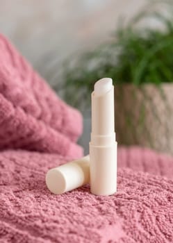 Opened lip balm stick on pink folded towel near green plant in bathroom,p, closeup, mockup. Lifestile scene with beauty product, everyday skincare routine