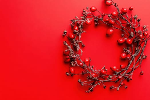 A circular wreath made of twigs and bright red berries sits against a vivid red backdrop creating a striking seasonal decoration.