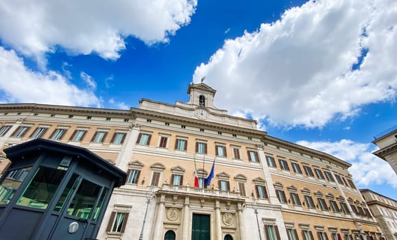 View of Palazzo Montecitorio in Rome, the seat of the Italian Chamber of Deputies, featuring Italian and European flags on the facade under a clear sky with clouds.
