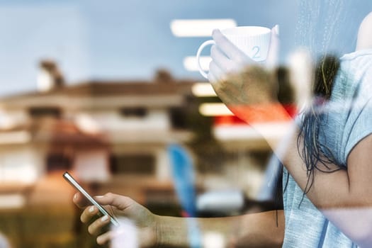 Woman is holding a smartphone and drinking coffee while standing by the window at home, her neighborhood is reflected in the glass