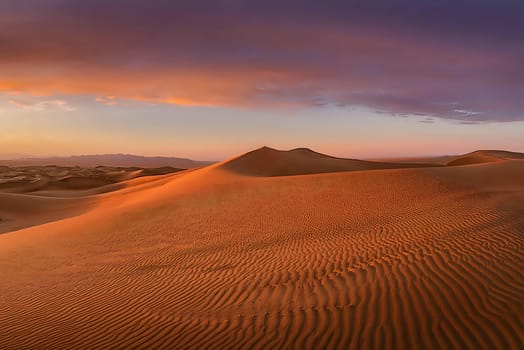 Expansive desert landscape showcasing rolling sand dunes under the warm light of the setting sun, with a gradient sky transitioning from soft oranges to deep purples