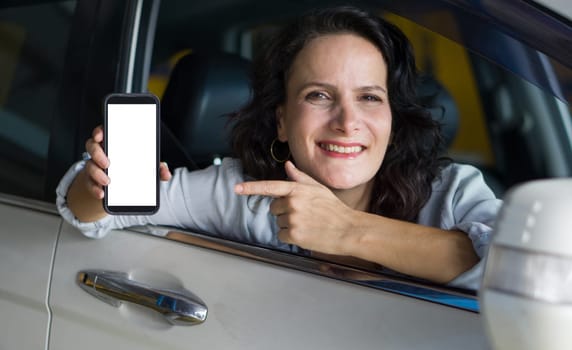Caucasian woman is sitting in the driver seat of a car with the window down. She is smiling and pointing at smartphone with a blank screen towards the camera.
