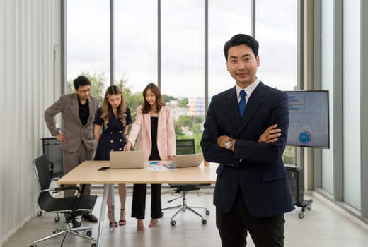 Asian businessman in a suit stand with arms crossed with a confident expression. The team is preparing for the meeting in the background. Concept of leadership.