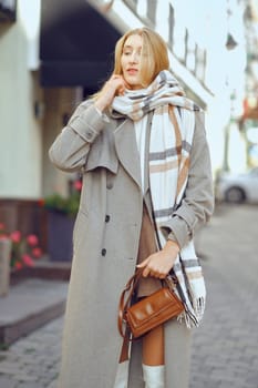 A young woman dressed in a chic overcoat and plaid scarf stands on a lively street, showcasing her fashionable urban look