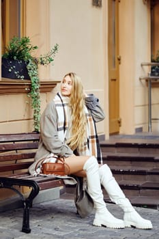 A woman with long hair relaxes on a bench dressed in a fashionable winter outfit. Her ensemble features a scarf and knee-high white boots, adding a chic flair to her serene moment.