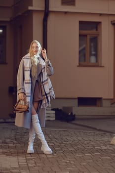 A young woman showcases her stylish ensemble on a cobblestone street in soft evening light. She wears a long coat and knee-high boots, holding a chic handbag with confidence and poise