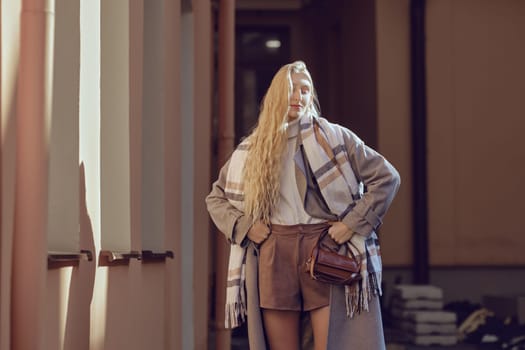 Stylish woman stands near the house under bright rays of sun, showcasing her fashionable outfit consisting of a cozy scarf, a fitted top, and tailored shorts, exuding confidence and style.