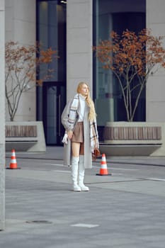 A fashionable woman with long hair wears a stylish outfit and stands in an urban area waiting for taxi. She exudes confidence as she poses next to orange traffic cones and modern architecture.