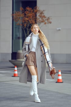 A confident woman walks through a modern urban area, showcasing an elegant combination of a stylish coat, shorts, and knee-high boots, accessorized with a chic plaid scarf on a bright day.