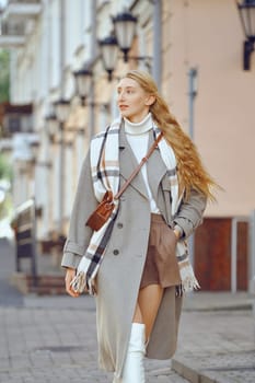 A woman in a gray overcoat and white sweater walks leisurely down a picturesque city street lined with lanterns. Her long hair flows in the breeze, and she carries a small bag.