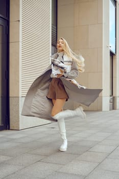 A young woman enjoys a carefree moment, twirling in her fashionable outfit of a coat and skirt. The urban backdrop features sleek architecture, enhancing the vibrant atmosphere of the day.