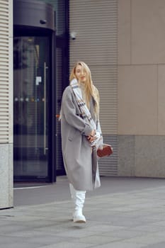 Dressed in a stylish long coat, a young woman strolls along a modern urban sidewalk, her hair gently swaying in the breeze. The contemporary architecture surrounds her as she enjoys a leisurely day.