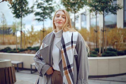 A young woman stands in a contemporary urban garden, showcasing her fashionable ensemble. She wears a cozy turtleneck and an oversized coat, exuding confidence amidst greenery and city architecture.