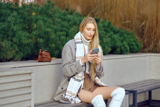 A young woman enjoys a leisurely moment in a park, sitting on a bench while checking her smartphone. Dressed in stylish fall attire, she exudes a relaxed vibe amidst the greenery.