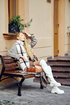 A woman with long hair sits gracefully on a bench, dressed in wool coat and a cozy scarf. She enjoys a moment of relaxation under the soft sunlight, surrounded by an urban setting.