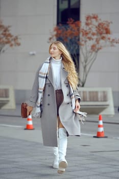A fashionable woman walks along a city street, wearing a stylish gray coat and knee-high white boots. She carries a small handbag and is surrounded by autumn foliage and construction barriers.