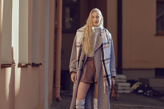 A young woman showcases her modern fashion sense, wearing a stylish coat and over-the-knee boots while standing against a backdrop of a warm-colored building on a sunny day.