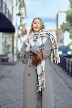 Cute woman standing on a moody city street, wearing a fashionable winter coat and a cozy scarf. She holds her accessories tightly as she enjoys the crisp air and vibrant atmosphere around her.
