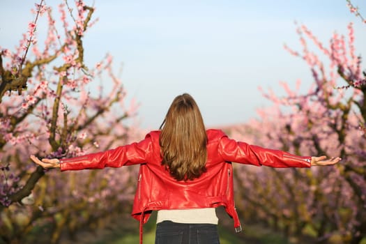 Back view portrait of a woman in red jacket outstretching arms in a field with pink flowers in spring season