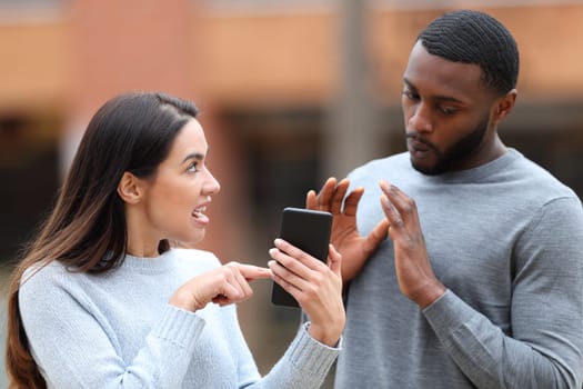 Angry woman scolding a shocked man showing cell phone content in the street