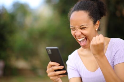 Excited black woman celebrating phone good news in a park