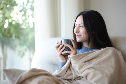 Relaxed asian woman looking through a window keeping warm and drinking covered with blanket coffee at home