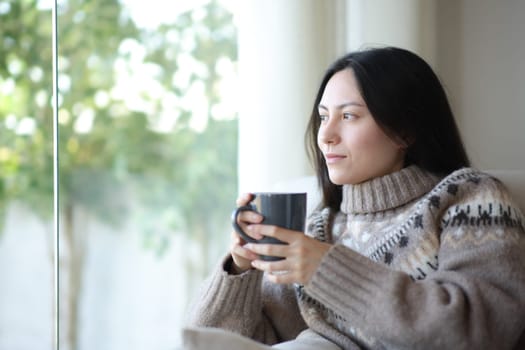 Serious asian woman relaxing in winter drinking coffee looking through a window at home