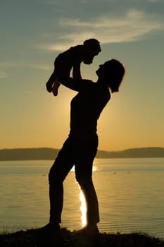 A beautiful silhouette of a parent lifting their joyful child against a serene sunset by the calm water