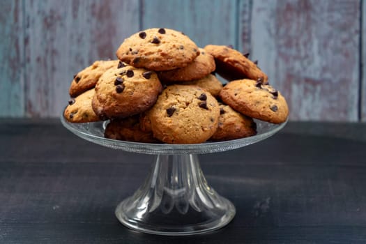A glass plate with a pile of chocolate chip cookies on it. The plate is on a wooden table