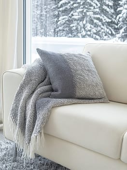 Comfortable white sofa with gray throw and pillow by a large window overlooking a snowy forest, creating a serene and inviting winter atmosphere.