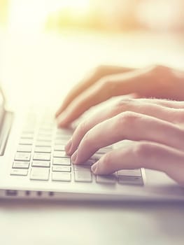 Close up view of hands typing on a laptop keyboard bathed in soft, warm light, representing modern work, communication, and technology use in a gentle atmosphere.