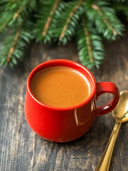 A vibrant red mug filled with creamy hot chocolate, topped with foam, sits on a rustic wooden surface beneath fresh pine branches, evoking cozy winter feelings.