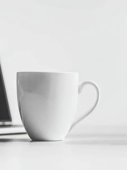 A simple white ceramic coffee mug sits on a light surface next to a blurred laptop, embodying a clean and modern workspace aesthetic.