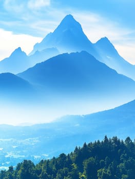 Stunning landscape of towering mountain peaks shrouded in misty blue hues, with forested foothills and a clear sky, capturing the serene beauty of nature.