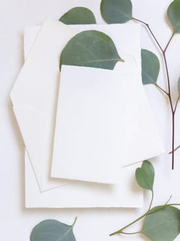 Card and envelope on a white table near green eucalyptus branches and leaves top view, Mockup. Minimal romantic flat lay with blank vertical card. Wedding and party stationery