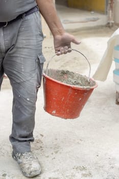 Craftsman hold in hand red bucket with cement, closeup detail of construction or rennovation process. Man in dirty working clothing and dusty hands