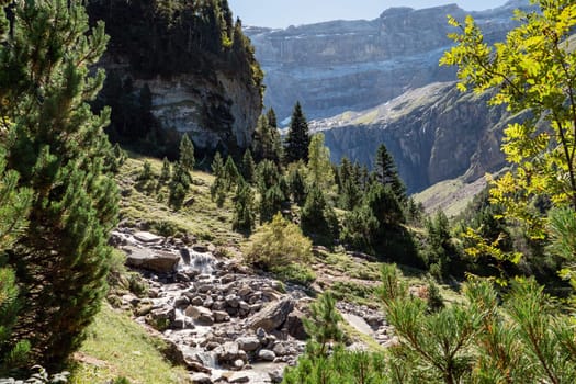 A mountain range with a stream running through it. The stream is surrounded by trees and rocks. The scene is peaceful and serene. Cirque de Gavarnie, France