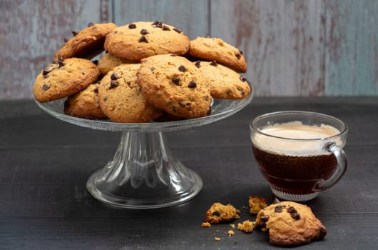 A glass of coffee and a plate of cookies. The cookies are chocolate chip and the coffee is black