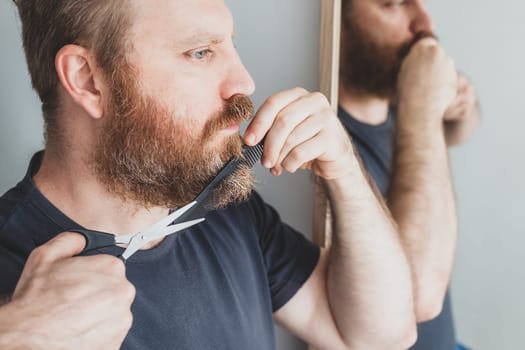 DIY Selfcare during quarantine isolation. Adult handsome caucasian man cutting moustache and beard himself personally with scissors at home. Coronavirus outbreak and people shutdown, stay home concept