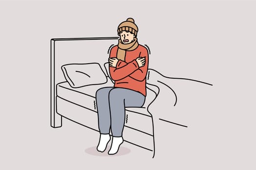 Freezing man is shaking sitting on bed and feeling chills and cold, dressed in hat and scarf. Freezing guy suffers due to lack of central heating system and poor immunity, located in bedroom