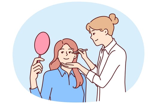 Woman beautician makes make-up of patient with mirror undergoing aesthetic treatment in medical clinic. Girl make-up beautician applies makeup on face of actress before filming movie or going on stage