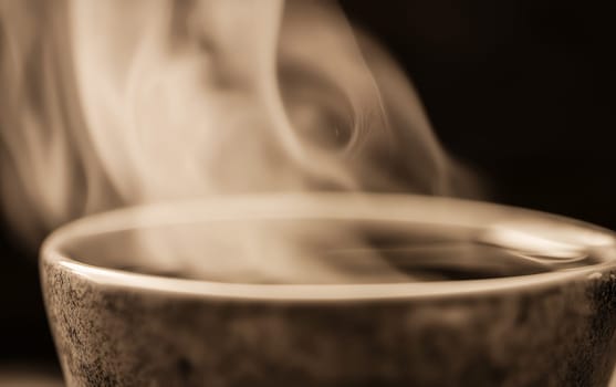 Close-up of a ceramic cup filled with hot coffee, wisps of steam rising gracefully, creating a cozy and inviting atmosphere in sepia tones.