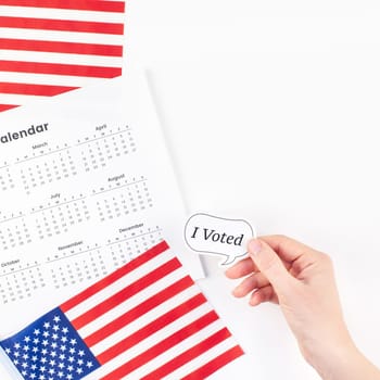 Creative top view flat lay of American flags for Elections Day with sticker I voted in hand with calendar copy space on white background in minimal style. Concept of patriotism and independence